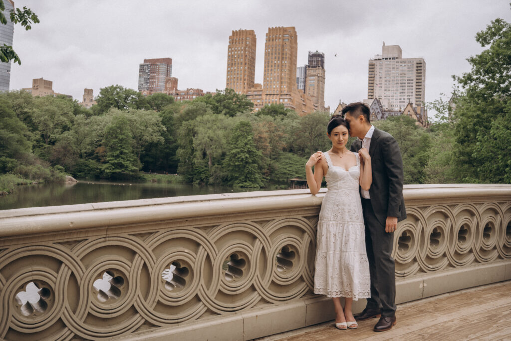 A groom kisses his bride standing on Bow Bridge in Central Park NYC, surrounded by lush greenery and the city skyline in the distance.